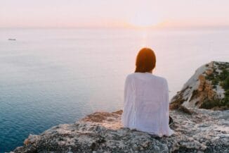 Mujer meditando en rocas frente al mar y el sol, simbolizando el despertar de la consciencia sobre el instinto de supervivencia