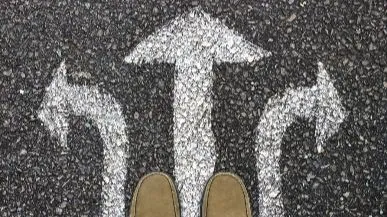 Pies de una persona frente a flechas pintadas en el suelo que indican múltiples direcciones (adelante, izquierda, derecha), simbolizando las elecciones del libre albedrío entre el ego y la perspectiva espiritual